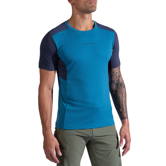 A man wearing the La Sportiva Ridge T-Shirt Men's in blue/navy and olive green pants stands against a white background, showing a tattoo on his left arm while looking slightly upward with arms relaxed at his sides.