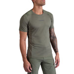 A man in an olive green La Sportiva Ridge T-Shirt and matching pants stands with his head turned slightly to the side, revealing a tattoo sleeve on his right arm. The shirt displays the La Sportiva logo on the chest.