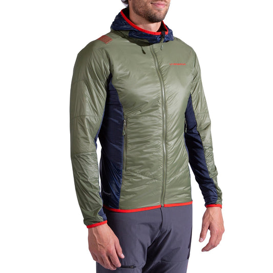 A man wears the La Sportiva Aequilibrium Lite Insulation Jacket Men's—an olive green and navy hooded jacket with red trim and a front zipper—paired with gray pants, standing slightly to the side with relaxed arms.