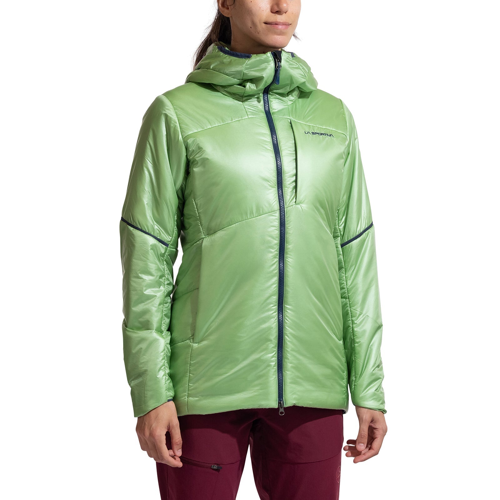A person in a shiny light green La Sportiva Supercouloir Primaloft Parka Women's—featuring a front zipper and zipped pockets—and maroon pants stands against a plain white background.