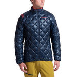 A man wearing the La Sportiva Lumina 100 Down Jacket Men's, a shiny dark blue insulated coat, and yellow pants stands against a plain white background.
