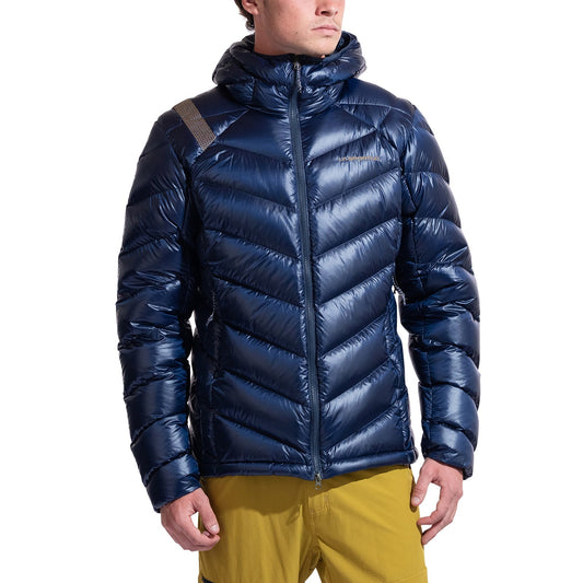 A man in a shiny, dark blue La Sportiva Lumina 300 Down Jacket with a hood and yellow pants stands slightly turned to the side against a white background.