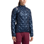 A woman in a La Sportiva Lumina 100 Down Jacket Women's in dark blue with a high collar and maroon pants stands against a white background.
