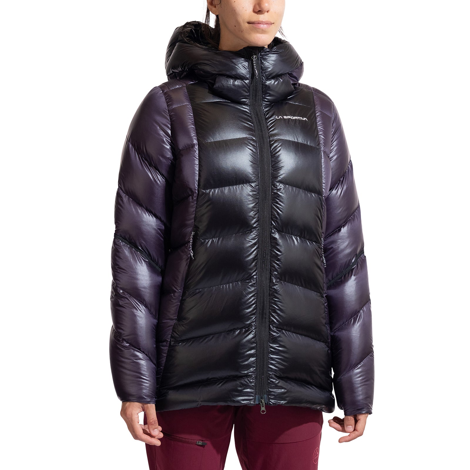 A person wears a shiny, black and dark purple La Sportiva Supercouloir Down Parka Women's with horizontal quilting, zipped up and paired with burgundy pants. This packable down jacket suits high-altitude climbing style against a white background.