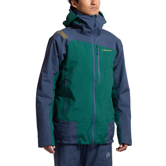 A man wearing the La Sportiva Alpine Guide GTX Performance Jacket Men's in green and dark blue, featuring zippered pockets and matching pants, stands against a white background.
