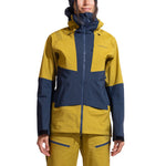 A woman stands against a white background wearing the La Sportiva Sender GTX Performance Jacket Women's in yellow and navy blue, featuring a high-collar hood, front zipper, and matching pants—ideal attire for ski touring. Her face is partially visible.