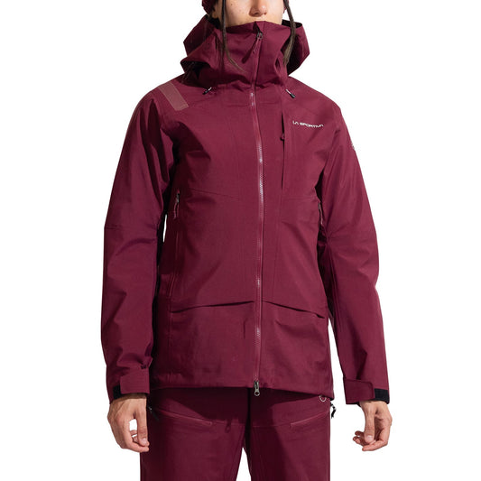 A woman faces forward wearing the La Sportiva Sender GTX Performance Jacket in maroon, featuring a high collar, hood, front zipper, and zippered pockets. The La Sportiva logo is on the chest—perfect for ski touring.