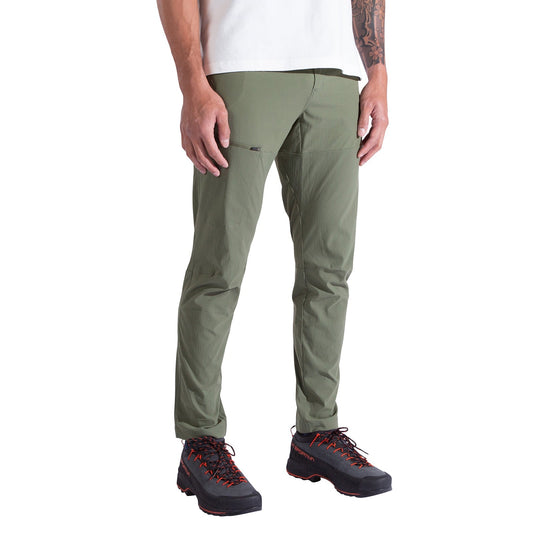 The image features the lower half of a person against a white background, wearing La Sportiva Machina Tech Pants Men's in olive green, paired with a white shirt and black hiking shoes with orange laces.