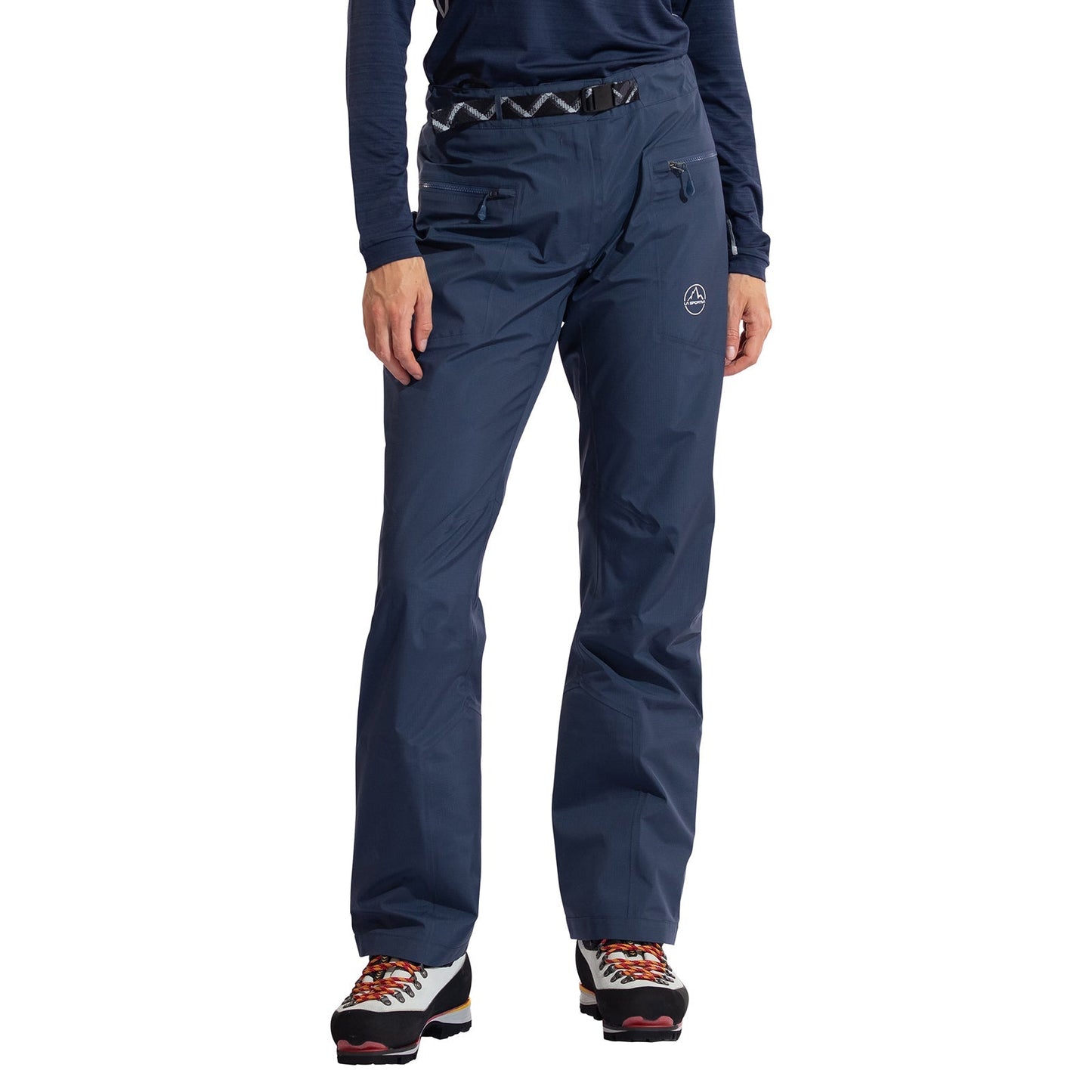 A woman stands against a white background wearing La Sportiva Alpine Guide GTX Performance Pants in navy blue, paired with a dark long-sleeve shirt and hiking boots. The waterproof pants have zippered pockets and an embroidered logo on the thigh.