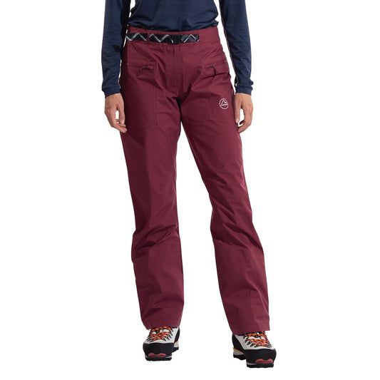 A woman stands against a white background wearing maroon La Sportiva Alpine Guide GTX Performance Pants Women's with GORE-TEX, a black belt, navy long-sleeve top, and hiking boots. The waterproof pants have zippered front pockets and a logo on the left thigh.