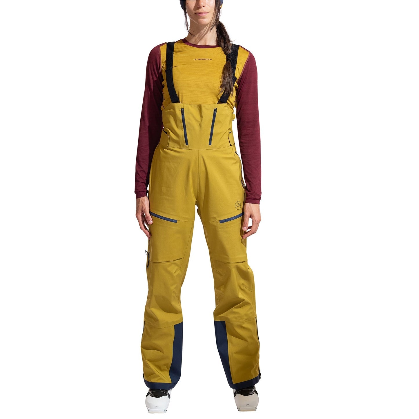 A person faces forward wearing La Sportiva Sender GTX Performance Bib Pants Women's in yellow with black straps, paired with a maroon long-sleeve shirt and white ski boots, set against a white background.