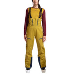 A person faces forward wearing La Sportiva Sender GTX Performance Bib Pants Women's in yellow with black straps, paired with a maroon long-sleeve shirt and white ski boots, set against a white background.