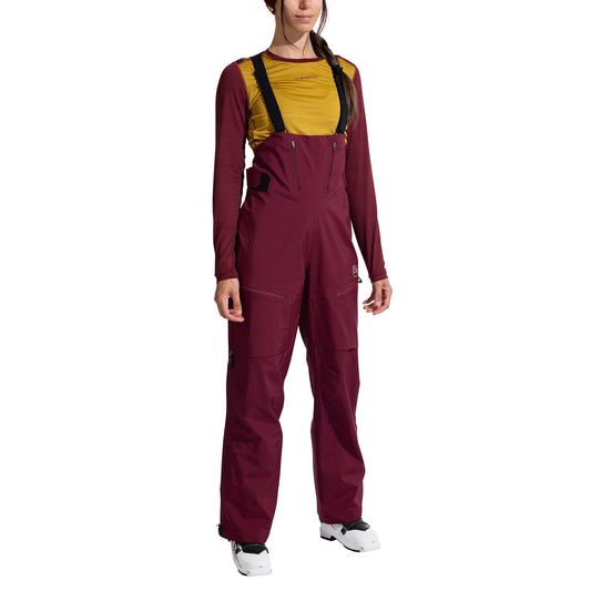 A woman models La Sportiva Sender GTX Performance Bib Pants Women’s in burgundy with black suspenders, paired with a mustard long-sleeve shirt and white ski boots. The front view is shown against a white background.