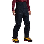 A person wears high-waisted black La Sportiva Supercouloir GTX Pro Pants Men's with pockets and zips, a maroon long-sleeve top, and yellow mountaineering boots against a white background—perfect pants for ice climbing.