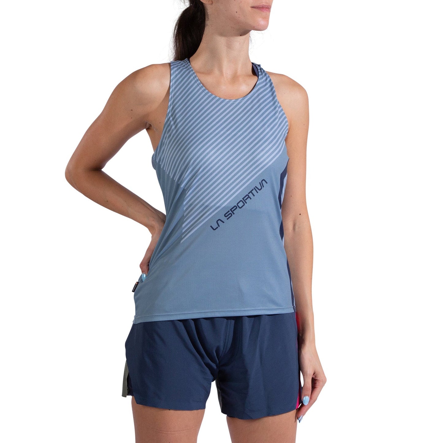 A woman wearing a La Sportiva Flow Tank Women's in light blue stripes, made from recycled polyester, and navy blue shorts stands with one hand on her hip against a white background.