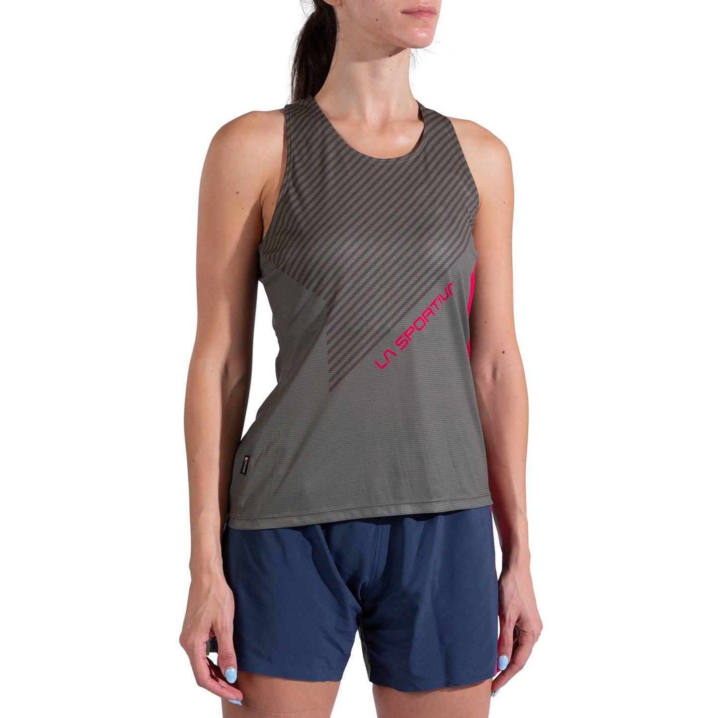 A woman stands against a white background wearing the La Sportiva Flow Tank Women's—an olive green, sleeveless top with diagonal stripes and a pink logo—paired with blue running shorts.