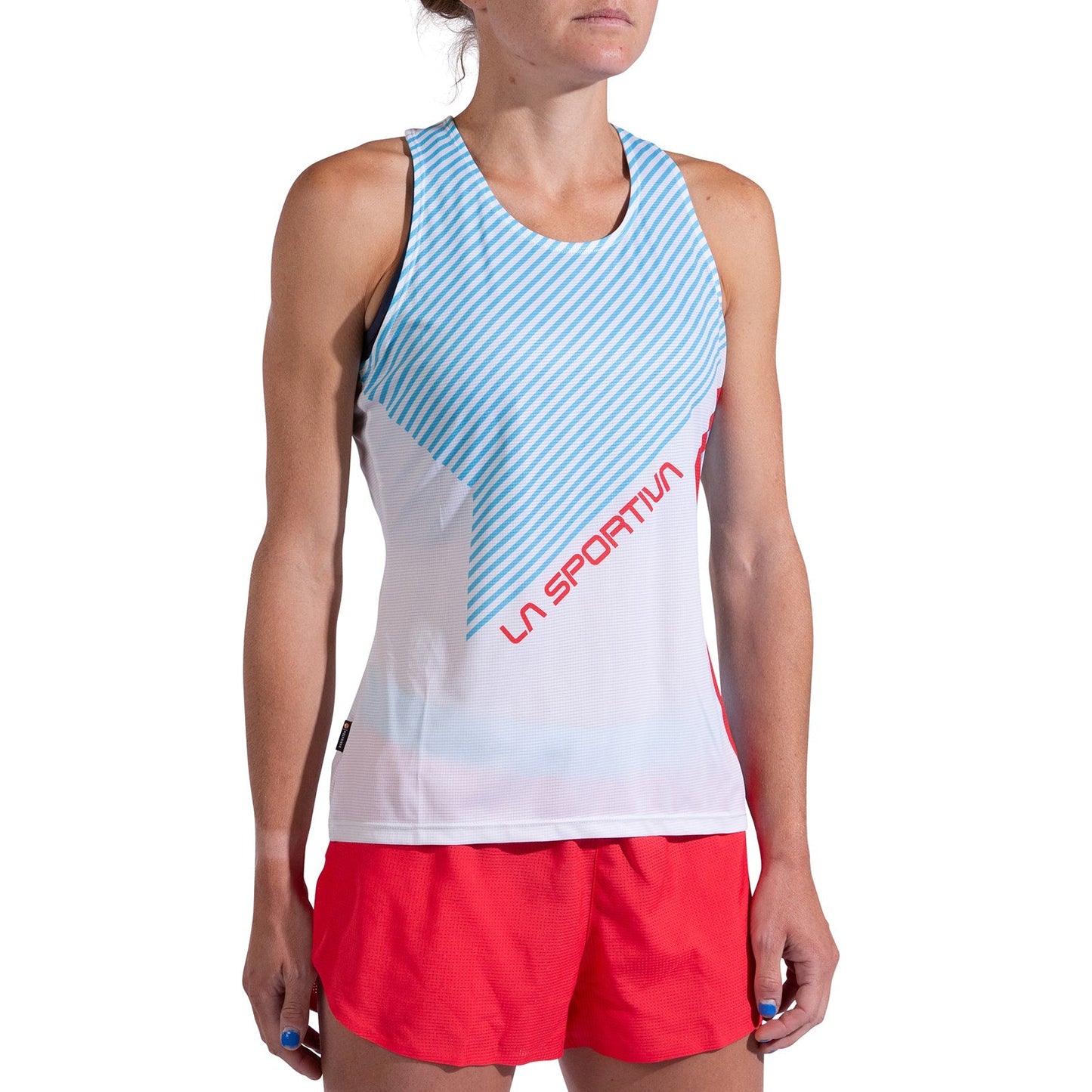 A woman stands with her arms relaxed by her sides against a white background, wearing a La Sportiva Flow Tank Women's in white and light blue stripes, made from recycled polyester, paired with red shorts.