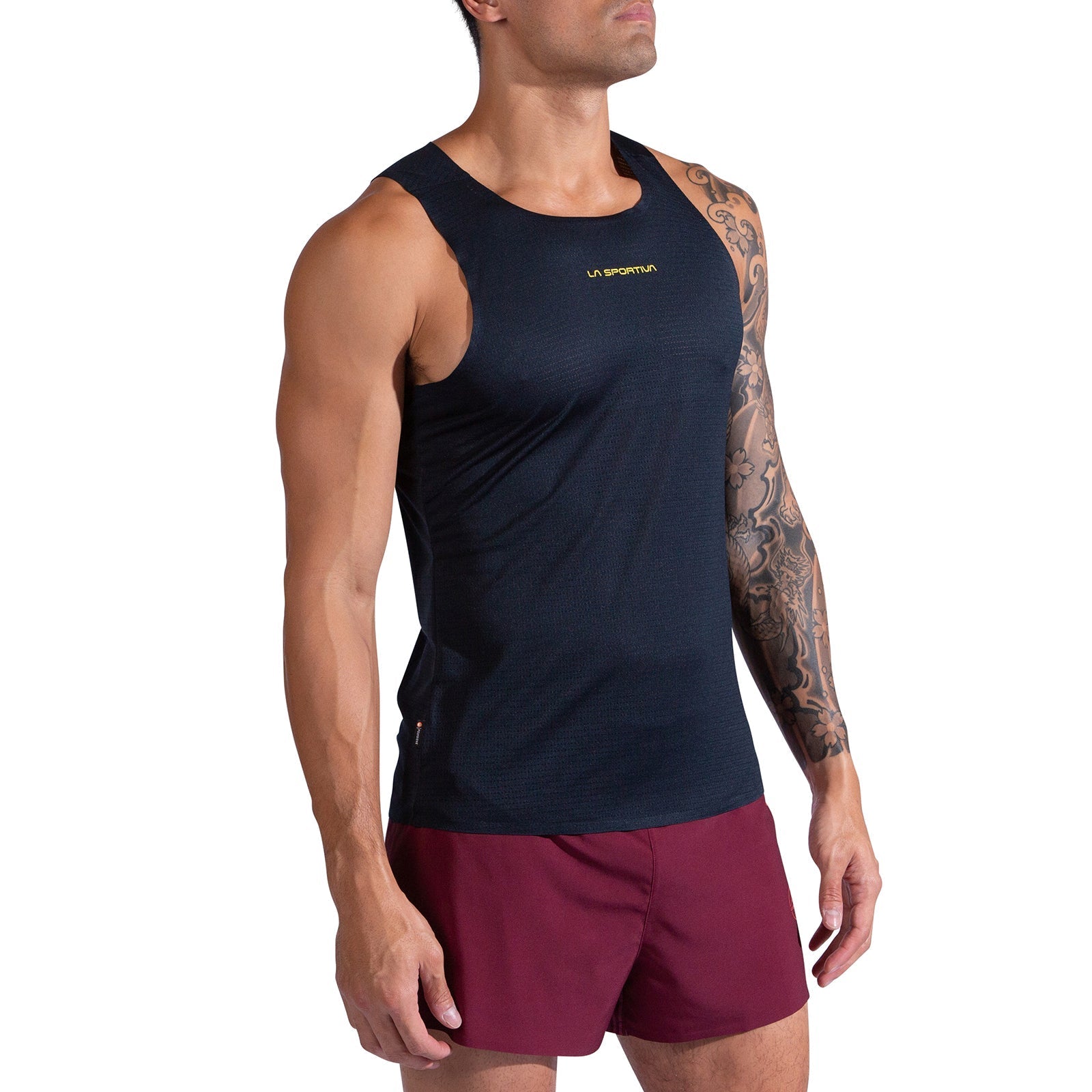 A man wearing the La Sportiva Pure Tank Men's black recycled polyester top and maroon shorts stands facing slightly left, showing a detailed tattoo sleeve on his right arm. The background is plain white.