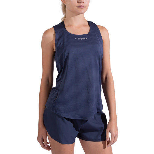 A woman wears the La Sportiva Pure Tank Women's in navy blue with matching shorts, standing against a white background. The sleeveless tank, made from recycled polyester, features the La Sportiva logo on the chest. Her head is slightly turned and arms relaxed.