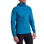 A man wears the La Sportiva Pocketshell Jacket Men's, a bright blue lightweight rain jacket with a front zipper and subtle logo detailing on the chest and sleeve, paired with black fitted pants, standing slightly turned to the side.