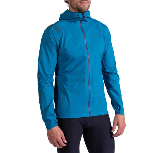 A man wears the La Sportiva Pocketshell Jacket Men's, a bright blue lightweight rain jacket with a front zipper and subtle logo detailing on the chest and sleeve, paired with black fitted pants, standing slightly turned to the side.
