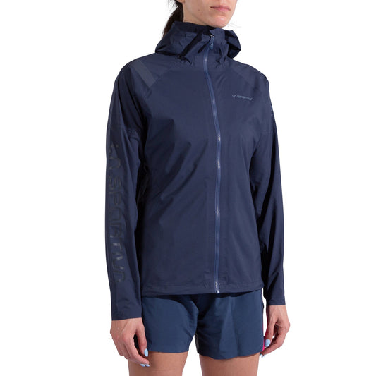 A woman wears the La Sportiva Pocketshell Jacket Women's in navy blue with a front zipper and matching shorts, standing against a plain white background.