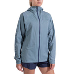 A woman stands against a plain white background wearing La Sportiva’s Pocketshell Jacket Women's in light blue, featuring the brand name on the left sleeve and chest, paired with dark blue shorts.
