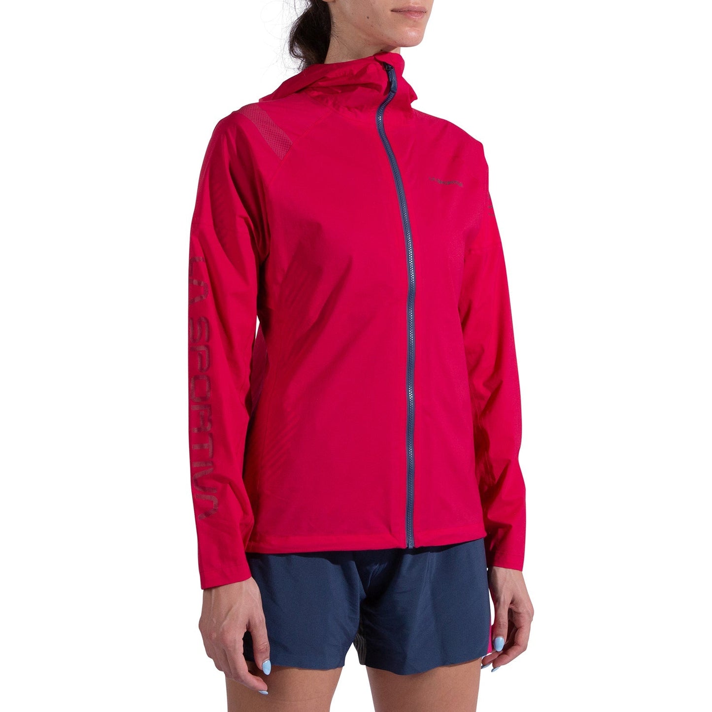 A person wears the La Sportiva Pocketshell Jacket Women's in bright pink with a hood and front zipper, featuring text on the left sleeve, paired with blue shorts. They stand against a white background, face partially out of frame.