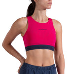 A woman stands with one hand on her hip, looking to the side, wearing the La Sportiva Captive Air Top Women's—a bright pink mountain running top with navy blue trim—paired with navy athletic shorts. This medium support bra is made from recycled fabric.