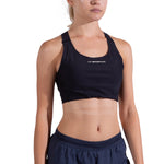 A woman in a black La Sportiva Pulse High Support Bra with crisscross straps and navy athletic shorts stands facing forward against a plain white background, her face partially out of frame.