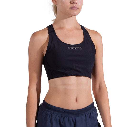 A woman in a black La Sportiva Pulse High Support Bra with crisscross straps and navy athletic shorts stands facing forward against a plain white background, her face partially out of frame.