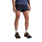 A person stands against a white background wearing La Sportiva Pure 3 Inch Shorts Men's, a navy blue shirt, white socks, and black trail running shoes with yellow and red accents. The person's face is not visible.