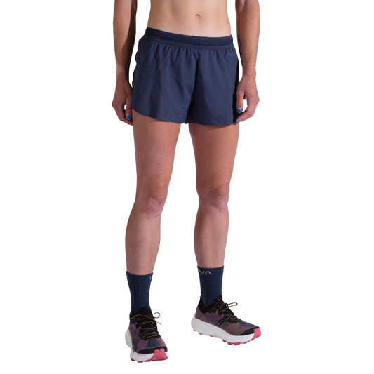 A person wears La Sportiva Pure 3 Inch Shorts Women's, dark crew socks, and gray and pink athletic shoes against a plain white background. The photo is cropped from the torso down, showing only the lower body.
