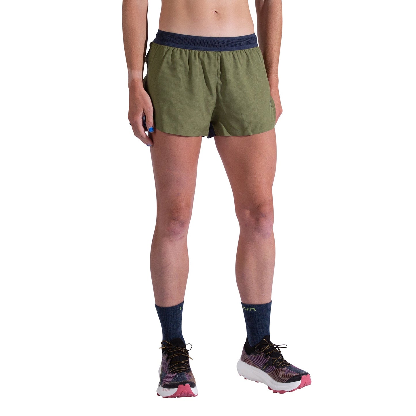 A person stands against a white background wearing La Sportiva Pure 3 Inch Shorts Women's in olive green with a dark waistband, navy socks, and white running shoes with pink soles. Only their torso and legs are partially visible.