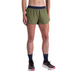 A person stands against a white background wearing La Sportiva Pure 3 Inch Shorts Women's in olive green with a dark waistband, navy socks, and white running shoes with pink soles. Only their torso and legs are partially visible.
