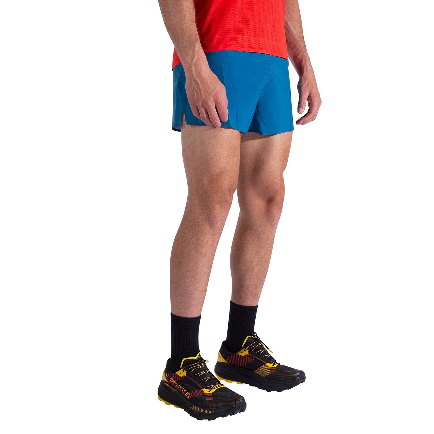 A man wears La Sportiva Flow 3 Inch Shorts Men's in blue, paired with a red shirt, black socks, and yellow-black trail running shoes. He stands against a white background with his upper body partially out of frame.