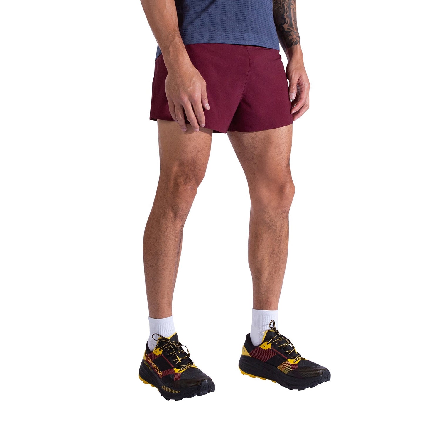 A person wearing a blue shirt, La Sportiva Flow 3 Inch Shorts Men's in maroon, white crew socks, and black running shoes with yellow accents stands against a white background.