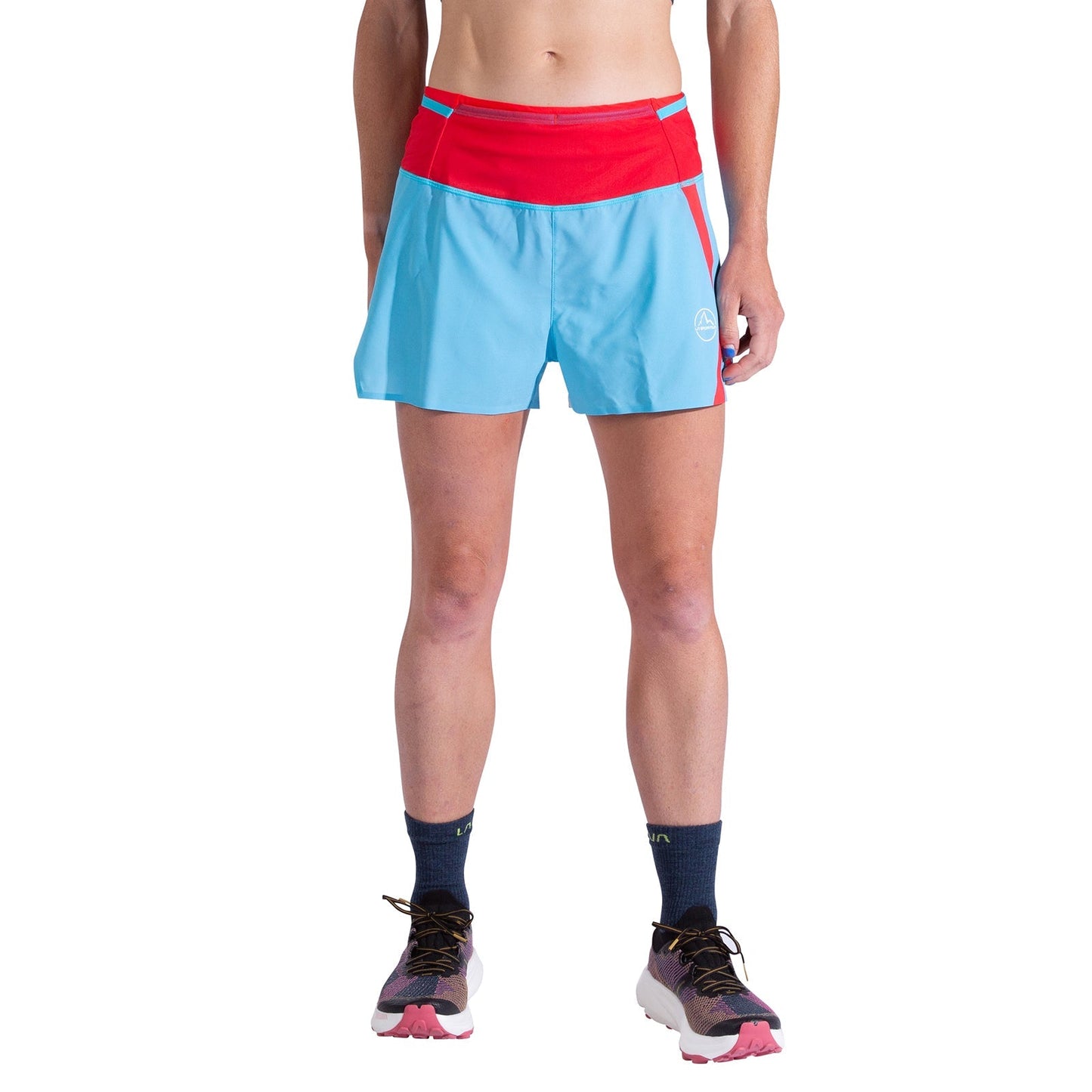 A person wears La Sportiva Flow 3 Inch Shorts Women's in blue with a red waistband and matching trim, paired with black socks and trail running shoes. Upper body and head are not visible; white background.