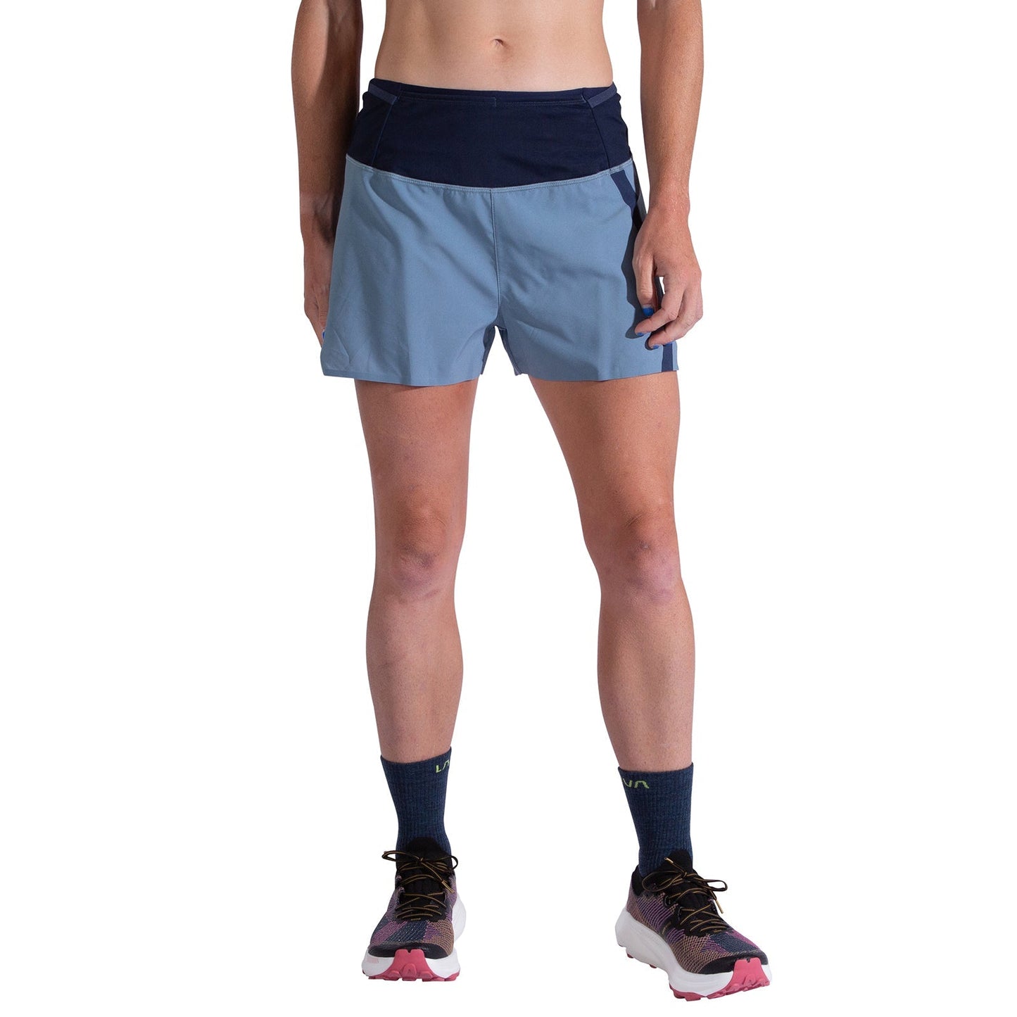 A person wears La Sportiva Flow 3 Inch Shorts Women's in light blue with a dark blue waistband, navy socks, and white running shoes with pink soles, standing against a white background. Their upper body is cropped out of the image.