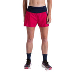 A woman wears La Sportiva Flow 3 Inch Shorts in bright red with a dark waistband, paired with navy socks and athletic shoes. She stands against a white background, visible from the chest down.