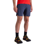 Only the lower half of a person is visible, showing La Sportiva Pure 7 Inch Shorts Men's in dark gray, paired with black socks and black-and-yellow running shoes against a white background.
