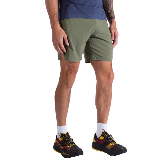 A person wears La Sportiva Pure 7 Inch Shorts Men's in olive green, a navy blue shirt, white socks, and black trail running shoes with yellow and red accents, standing against a white background.