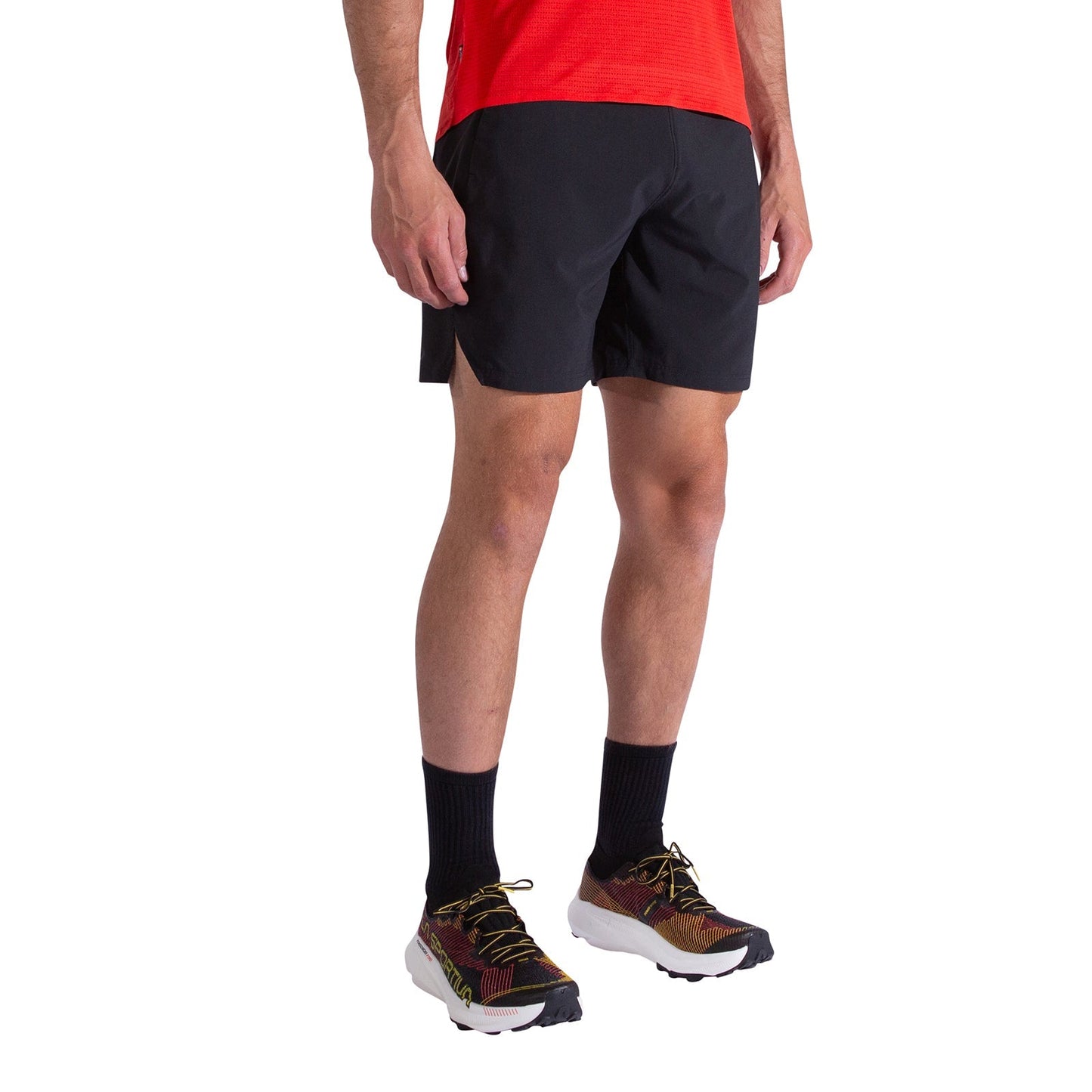 A person wearing La Sportiva Pure 7 Inch Shorts Men's in black, along with a red shirt, black crew socks, and multicolored running shoes stands against a white background; only the lower half of the body is visible.