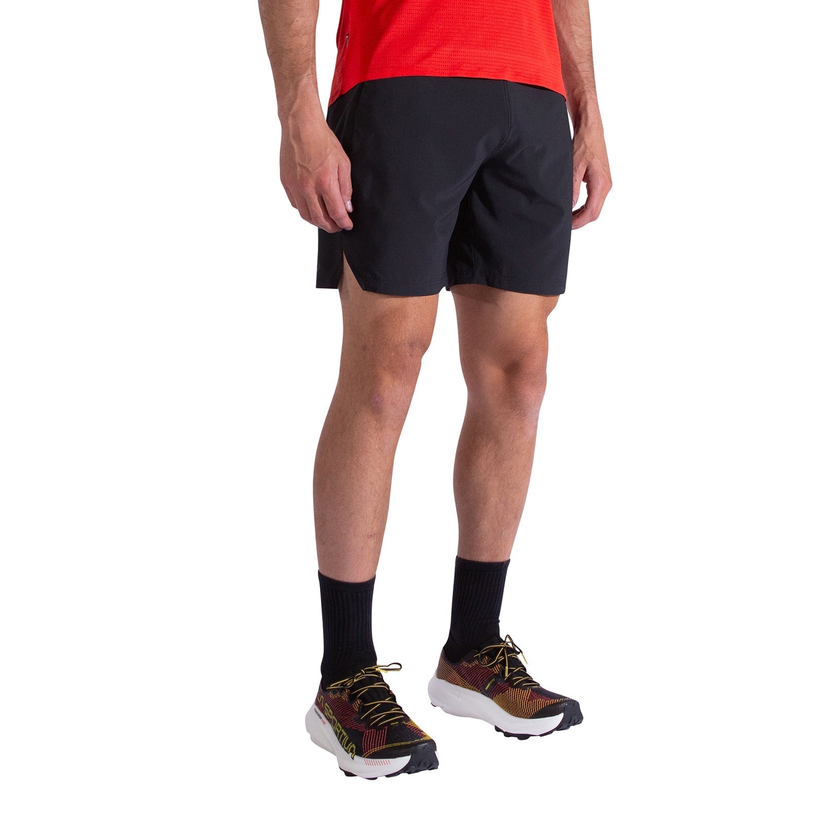 A person wearing La Sportiva Pure 7 Inch Shorts Men's in black, along with a red shirt, black crew socks, and multicolored running shoes stands against a white background; only the lower half of the body is visible.