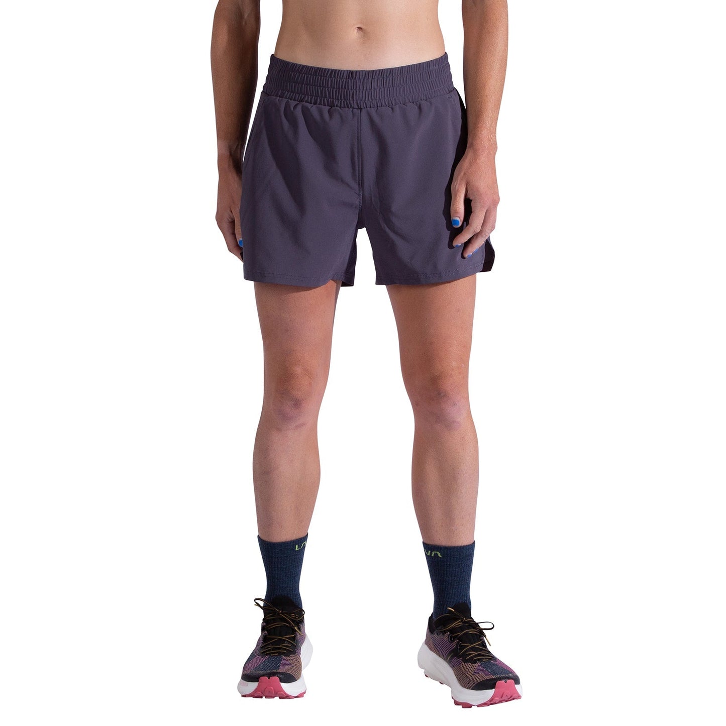 A person is wearing La Sportiva Pure 4 Inch Shorts Women's in dark purple, navy socks, and running shoes with pink and white accents, standing against a plain white background with relaxed hands by their sides.