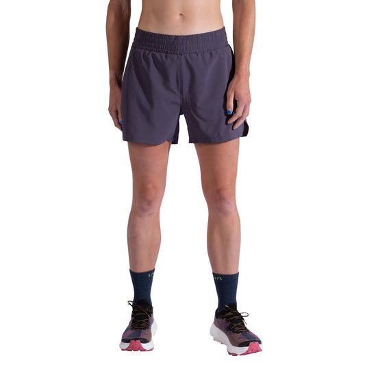 A person is wearing La Sportiva Pure 4 Inch Shorts Women's in dark purple, navy socks, and running shoes with pink and white accents, standing against a plain white background with relaxed hands by their sides.