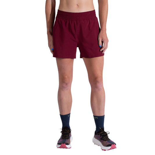 A person models La Sportiva Pure 4 Inch Shorts Women's in maroon, paired with dark navy socks and white running shoes with pink soles. Only their lower torso and legs are visible against a white background.