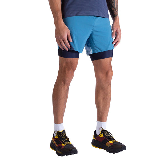 A person wearing La Sportiva Flow 5 Inch Shorts Men's in blue, a matching T-shirt, white socks, and black running shoes with yellow and red accents stands against a white background.
