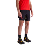 A person wearing a red shirt, La Sportiva Flow 5 Inch Shorts Men's (black with yellow accents), black socks, and black running shoes with yellow and red details, standing against a white background.