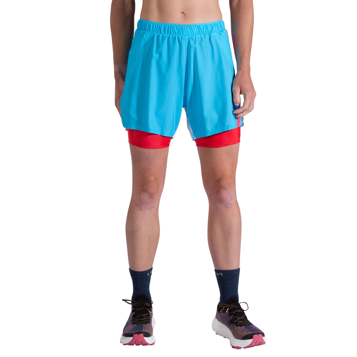 A woman stands in La Sportiva Flow 5 Inch Shorts, featuring navy socks and red compression shorts underneath. Only her lower torso and legs are shown against a white background; breathable stretch fabric ensures comfort.